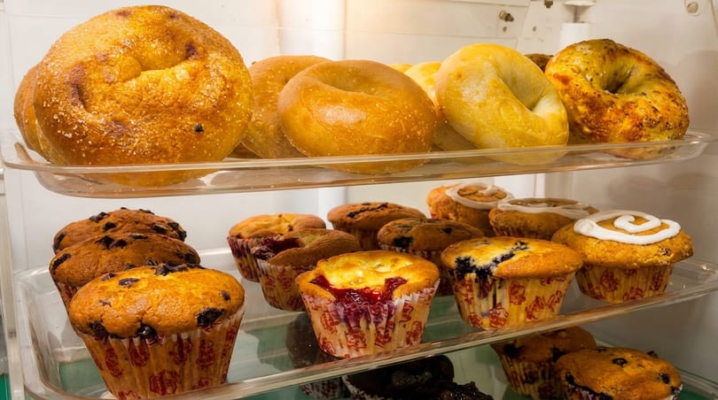 Freshly baked bagels, blueberry muffins, and frosted sweet rolls on clear display shelves.