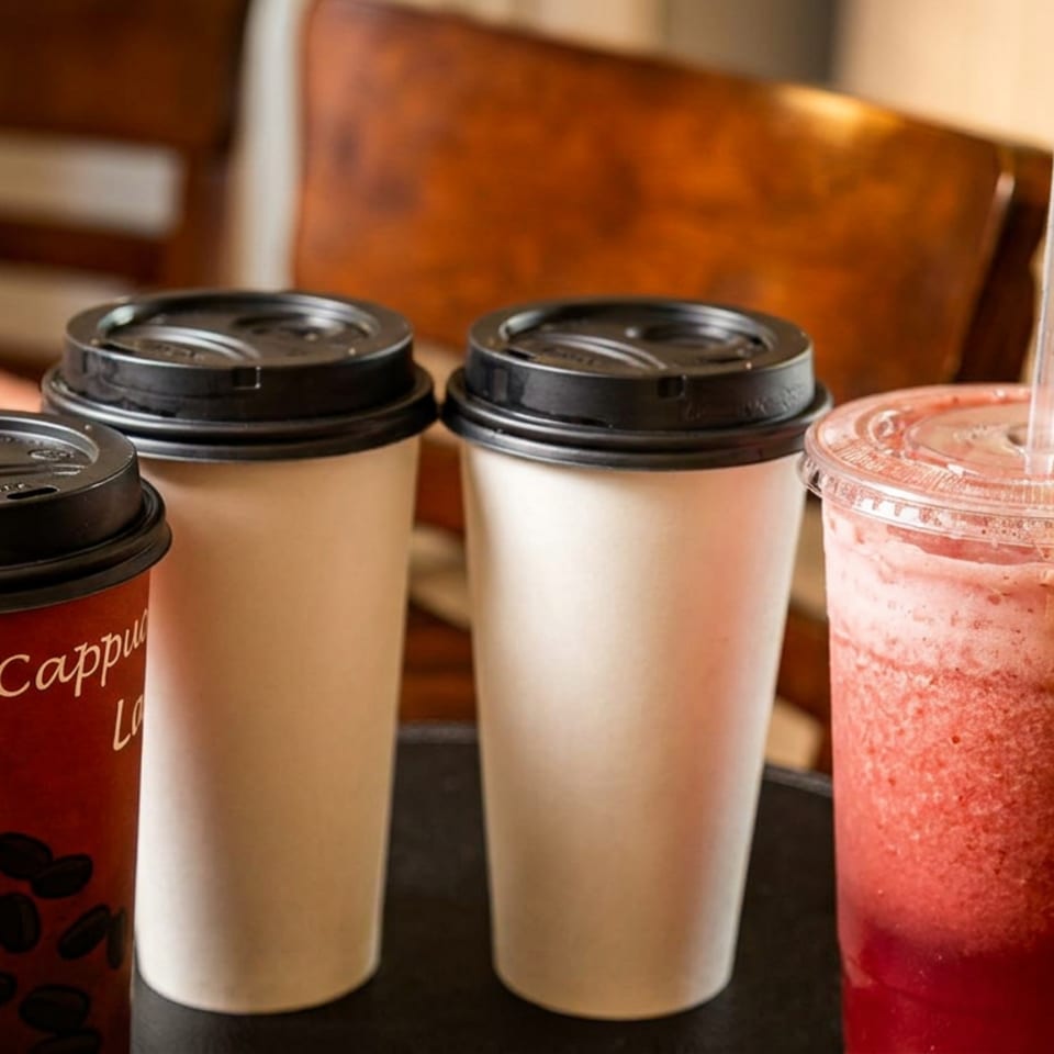 Three coffee cups, one red 'Cappuccino', and a strawberry smoothie with a straw on a dark surface.