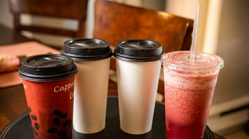Three coffee cups, one red 'Cappuccino', and a strawberry smoothie with a straw on a dark surface.
