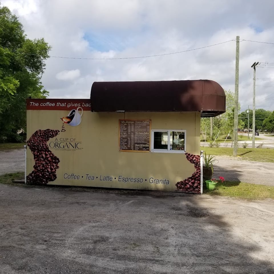A Cup of Organic coffee stand with coffee bean graphic, menu, and drive-thru window in a gravel lot.