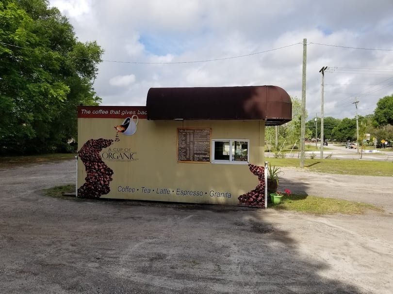 A Cup of Organic coffee stand with coffee bean graphic, menu, and drive-thru window in a gravel lot.