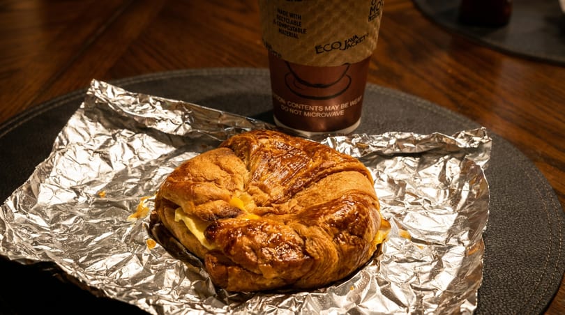 Golden croissant sandwich on foil with a disposable coffee cup on a dark placemat.
