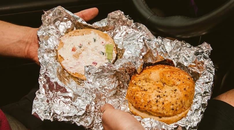Two bagels, one with cream cheese and celery, wrapped in foil and held in a car.